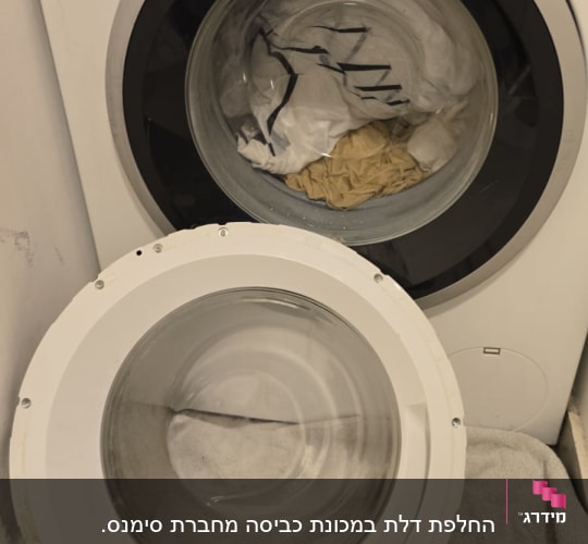 מכונת כביסה פתוחה עם בגדים בפנים ודלת מונחת בצד